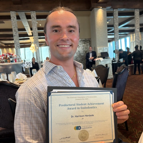 A close-up shot of Dr. Harrison Herdade smiling while holding his framed "Predoctoral Student Achievement Award in Endodontics" certificate inside an event venue.