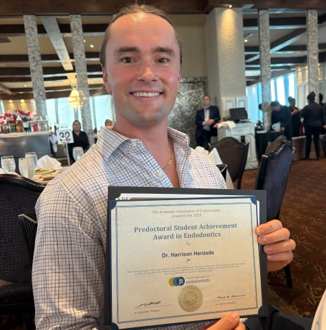 A centered photo of Dr. Harrison Herdade holding an award certificate from the American Association of Endodontists in a formal indoor event space.