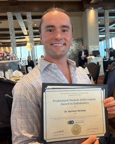A smiling man in a plaid collared shirt holding up an award certificate titled "Predoctoral Student Achievement Award in Endodontics" presented to Dr. Harrison Herdade by the American Association of Endodontists, set in a banquet dining room.